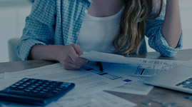 #PraCegoVer Texto: BOLETOS. Imagem: mulher de camisa xadrez está sentada em frente à mesa com notebook aberto, calculadora e vários boletos.